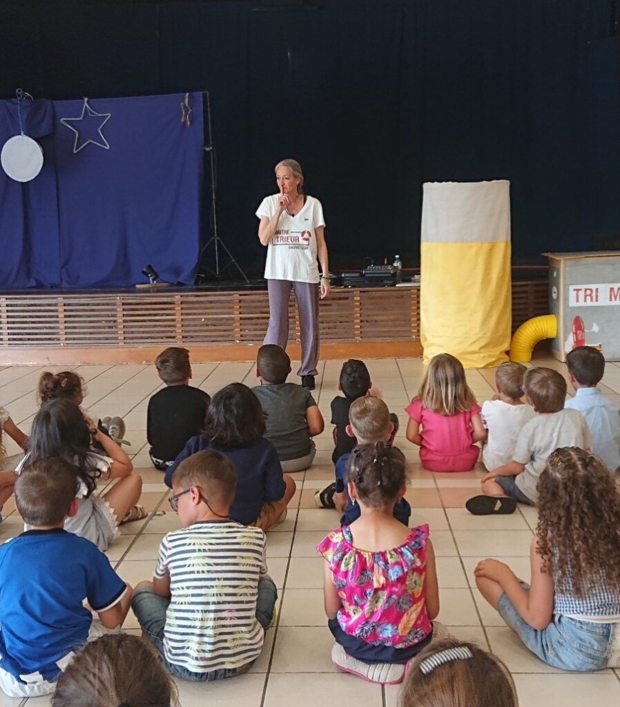 Photo du spectacle copains, copine, dans une salle avec le décor et la machine trimalin. Les enfant de dos écoutent attentivementle Maitre Trieur Sauveteur.